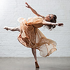 A female dancer dressed in a light, flowy dress outstretches her left leg while standing on her right tiptoes.