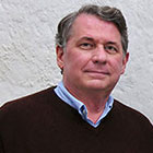 Phillip Setzer posses for a photo wearing a collared shirt and sweater in front of a stucco wall.