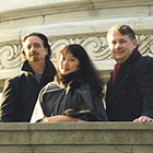 David Finckel, Wu Han, and Philip Setzer stand along an ornate wall and railing wearing coats.