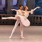 A male dancer holds up a female dancer while she stands and extends her right leg at a 90-degree angle behind her.
