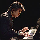 A musician dressed in a suit sits and plays a composition on the piano.