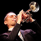 A man wearing a suit closes his eyes as he plays a trumpet pointed upward.