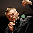 Karabits, eyes closed and dressed in a dark suit, raises his baton and left hand.