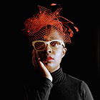The singer, wearing thick-rimmed glasses and a decorative netted hat, holds her right hand to her cheek.