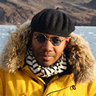 Miller, wearing a fur hooded coat and dark glasses, stands in front of the Arctic Sea.