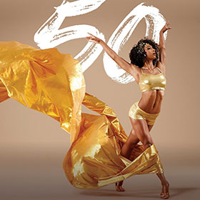 A female dancer raises her arms in the air as a plume of golden fabric flows in her wake.
