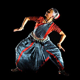 A dancer wearing traditional Indian dress stands with her arms and legs at shoulder width and bent at the elbows and knees while she looks up to the side.