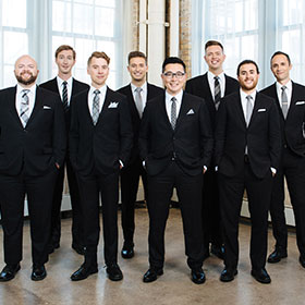 The eight members of Cantus stand in a row wearing black suits.