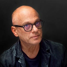 A portrait of a bald man wearing bold glasses and a leather jacket.