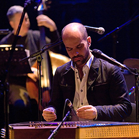 A balding man looks down while he plays a horizontal stringed instrument called a santur.