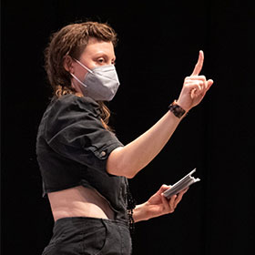 A woman wearing a face mask stands and holds index cards in her left hand and her right pointer finger straight up. 