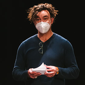 A man with curly hair holds index cards in both hands as he looks straight at the camera.