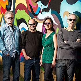 Quartet members wearing sunglasses stand against a mural wall.