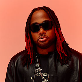 A portrait of Leon Thomas wearing a leather jacket, large sunglasses, and a metal chain around his neck.