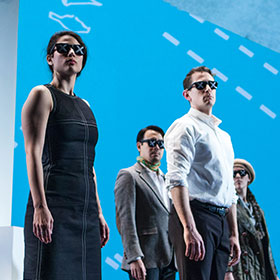Actors walk across the stage wearing sunglasses while charts and maps are projected onto them.