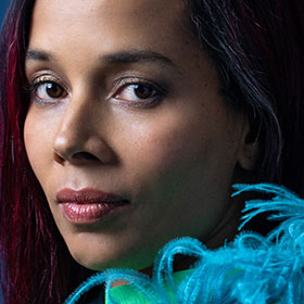 A woman of color wears a shawl with a feathery fringe as she turns her head for a portrait.