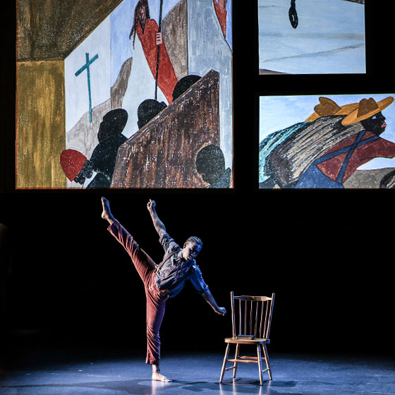 A dancer balances against a chair and extends an arm and a leg into the air. Projected behind him are three paintings.